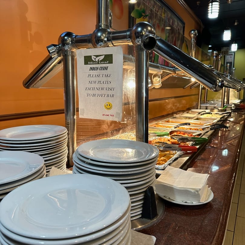 A Mint Leaf Indian Cuisine buffet line with stacks of plates, napkins, and hot food.