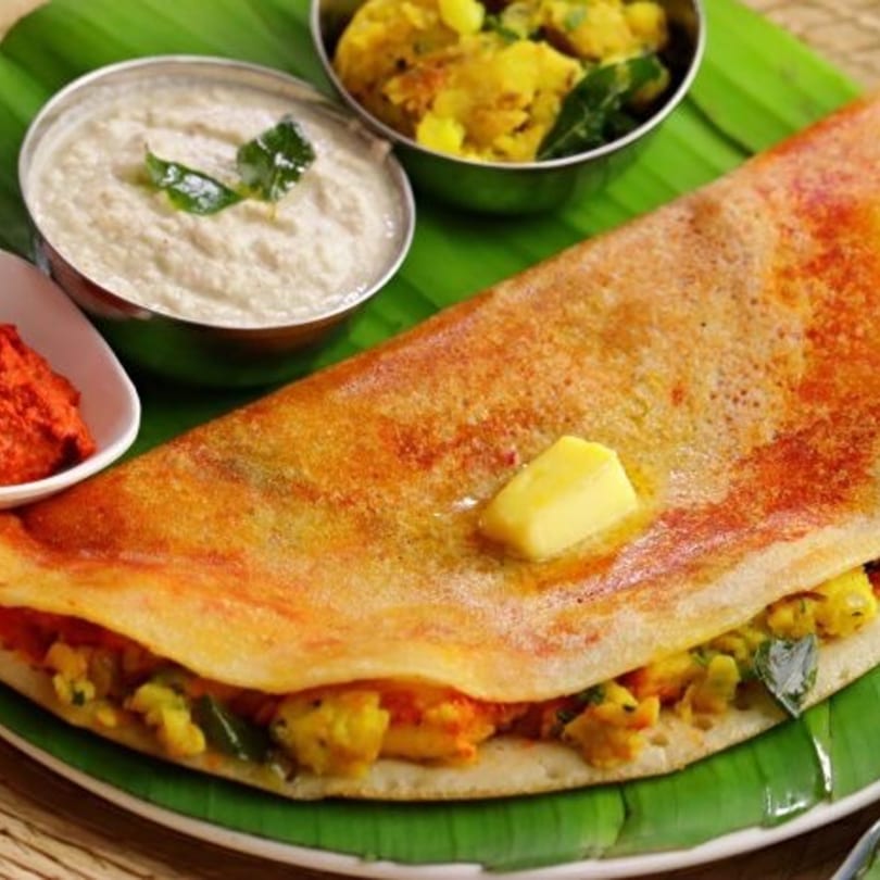A golden-brown dosa with potato filling, butter, and chutneys on a banana leaf.