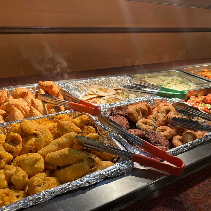 Steaming hot Indian buffet with samosas, pakoras, and other appetizers in foil-lined trays.