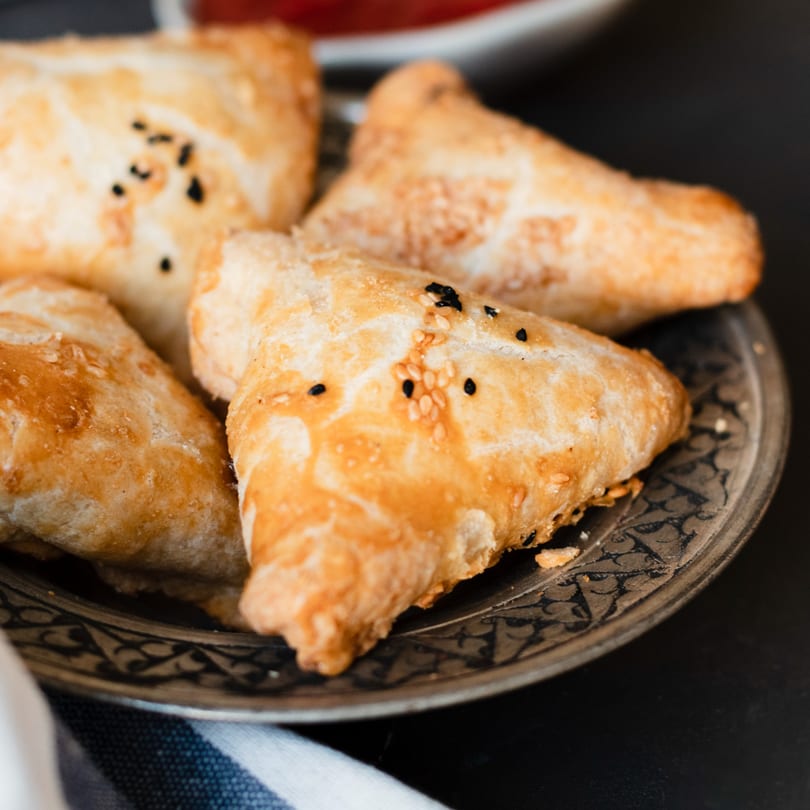 Golden-brown Indian samosas, sprinkled with sesame and nigella seeds, on an ornate metal plate.